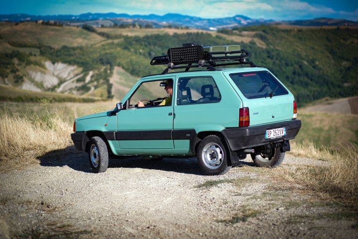 A mint green vintage hatchback car with roof rack parked on a scenic hillside.