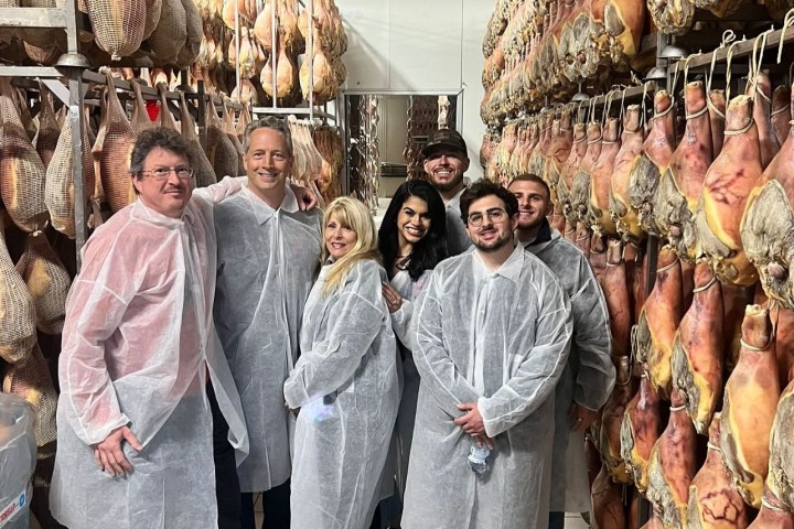 People in white coats in a room lined with hanging cured meats on racks.