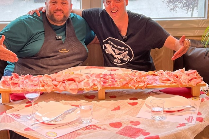 Two men smiling, sitting at a table with a large tray of sliced meats.
