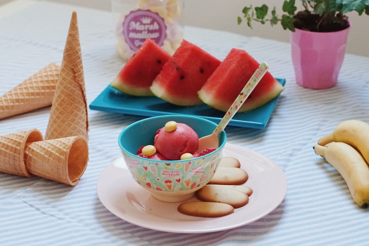 Ice cream and cookies on a plate, cones, watermelon slices, bananas, and a plant on a striped tablecloth.